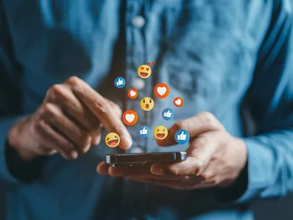 Person tippt auf Smartphone, während bunte Social-Media-Emojis wie Likes und Herzen darüber schweben