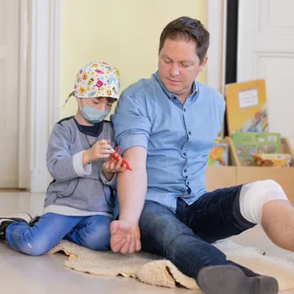 Junge mit OP-Haube und Spielzeug-Spritze „behandelt“ erwachsenen Patienten – emotionales Kampagnenmotiv für den Beruf des Anästhesietechnischen Assistenten (ATA) im Klinikverbund Allgäu.