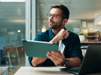 Ein Webentwickler sitzt in einem modernen Büro vor Laptop und Tablet, schaut nachdenklich zur Seite und reflektiert über digitale Lösungen. Die Szene symbolisiert kreative Denkprozesse, agiles Arbeiten und technologiegestützte Markenentwicklung in einer Agenturumgebung.