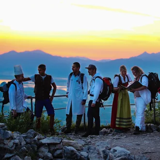 Gruppe von Klinikmitarbeitenden verschiedener Berufsgruppen auf einem Berggipfel bei Sonnenuntergang – starke Metapher für interdisziplinäres Teamwork und berufliches Wachstum bei der Fachklinik Allgäu.