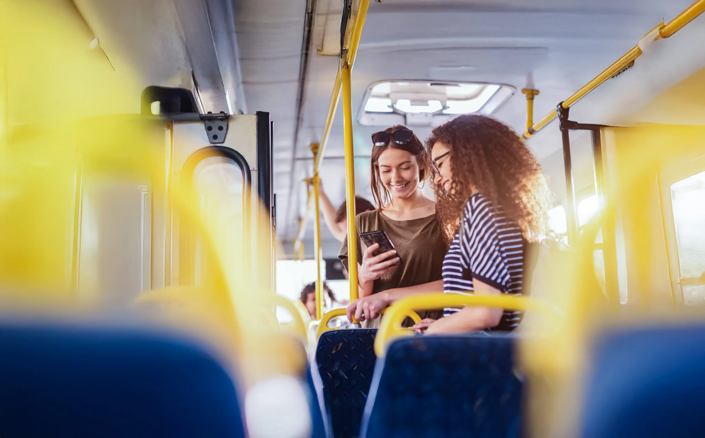 Zwei junge Frauen stehen in einem Bus und schauen gemeinsam auf ein Smartphone; sonniger Tag, gelbe Haltestangen und Sitzreihen im Vordergrund