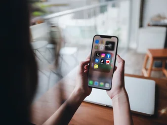 Person hält Smartphone mit geöffnetem Social-Media-Ordner, der Icons von Instagram, Facebook, LinkedIn, TikTok, YouTube, Snapchat und X zeigt – Darstellung digitaler Vernetzung und mobiler Mediennutzung