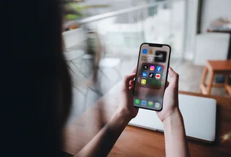 Person hält Smartphone mit geöffnetem Social-Media-Ordner, der Icons von Instagram, Facebook, LinkedIn, TikTok, YouTube, Snapchat und X zeigt – Darstellung digitaler Vernetzung und mobiler Mediennutzung