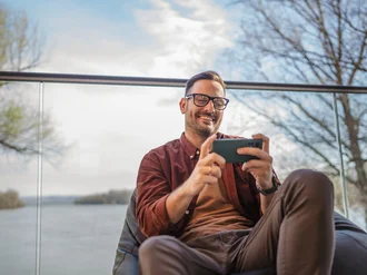 Lächelnder Mann mit Brille sitzt entspannt auf einer Terrasse am Wasser und nutzt sein Smartphone – steht für digitale Freizeitgestaltung, mobiles Streaming und moderne Work-Life-Balance