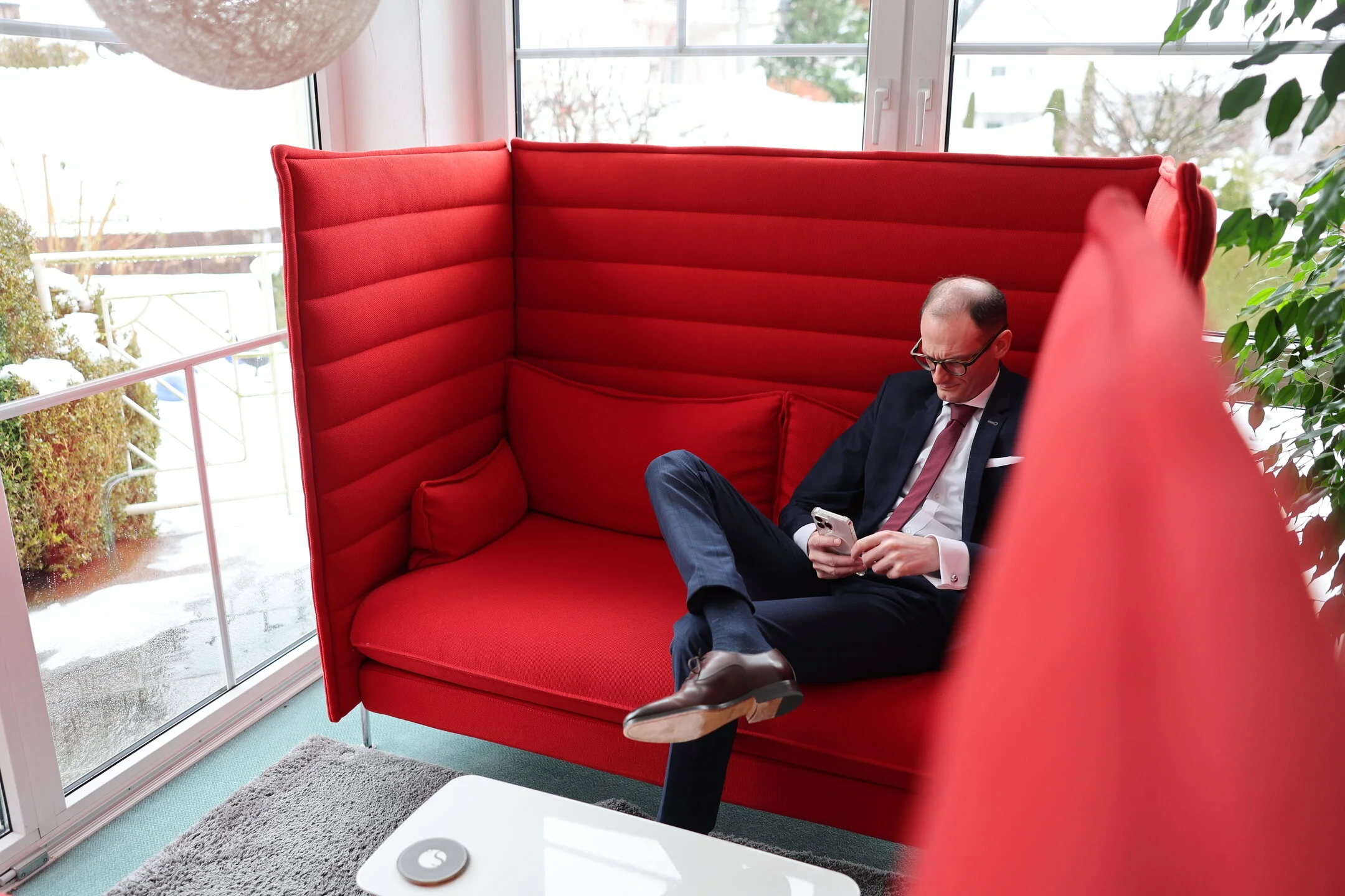 Denkströme Geschäftsführer im Anzug sitzt entspannt in moderner roter Lounge-Sitznische und nutzt Smartphone – Rückzugsort für konzentriertes Arbeiten im Büro.